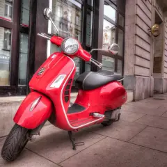 Czerwona karta motorowerowa Vespa zaparkowana na chodniku przed budynkiem.