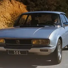 Srebrny Peugeot 504 z lat 70. jedzie po krętej drodze wśród wzgórz.