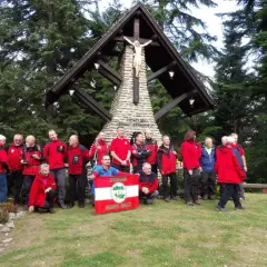 Grupa turystów w czerwonych kurtkach pozuje przy kamiennym krzyżu w Beskidach. W tle las i drzewa.