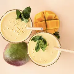 Zwei Gläser cremiger, veganer Mango Lassi, garniert mit Minze, neben einer ganzen und einer gewürfelten Mango.