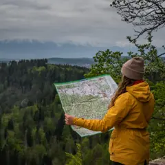 Kobieta w żółtej kurtce analizuje mapę, planując wędrówkę po malowniczej Słowackiej Szwajcarii. W tle rozciąga się zielony las i mgliste góry.