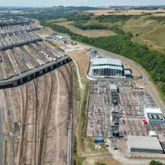 Rozległa infrastruktura kolejowa, gdzie pociąg Eurotunelu przygotowuje się do podróży. W tle wzgórza i autostrada.