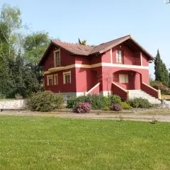 Una casa típica cántabra, de color rojo intenso con detalles amarillos, rodeada de vegetación y un césped verde.