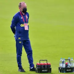Mężczyzna w niebieskim dresie i masce na twarzy, z napisem "ML" na nogawce, stoi na boisku. To kitman, który dba o zawodników.