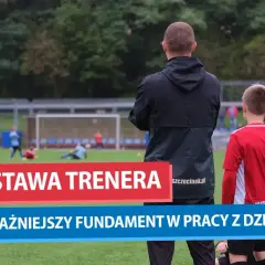Trener motywacyjny obserwuje młodych piłkarzy na boisku. Jego postawa to fundament pracy z dziećmi.
