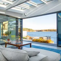 Soggiorno luminoso con vista lago, divano grigio e tavolino in legno. L'ampia vetrata e il soffitto trasparente creano un perfetto stile coastal.