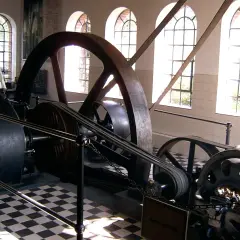 Eine historische Dampfmaschine, Symbol der Industrialisierung, mit großen Schwungrädern und Riemenantrieb. Ein Mann in Arbeitskleidung steht daneben.