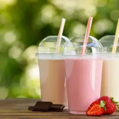 Trzy kolorowe szejki w plastikowych kubkach z rurkami, obok truskawki, banany i kawałki czekolady.