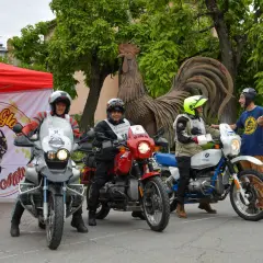 Tre motociclisti pronti per un percorso eroica in moto, con bandiera italiana e un grande gallo di metallo sullo sfondo.