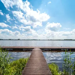 Drewniane molo prowadzi na spokojne jezioro. W oddali widać wiatraki, a nad głową błękitne niebo z białymi chmurami. Idealne miejsce na skalno-wypoczynkowy relaks.