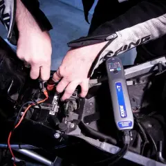 Mani che collegano un caricabatteria Exide alla batteria di una moto. Scopri come caricare batteria moto senza caricabatteria.