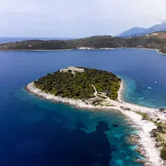Isla con un castillo en Himare, Albania, rodeada de aguas azules y vegetación exuberante. Un muelle y barcos anclados completan la vista.