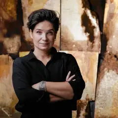 Agnieszka Smoczyńska, reżyserka o krótkich, ciemnych włosach, w czarnej koszuli, z rękami skrzyżowanymi na piersi, na tle abstrakcyjnych, złotobrązowych płócien.