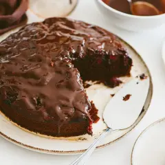 Pyszny brownie z malinami, polane błyszczącą czekoladą, czeka na talerzu. Obok stoi łopatka do ciasta.