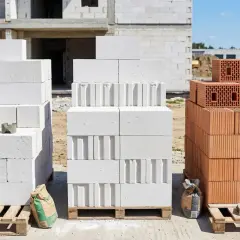 Porównanie materiałów budowlanych: biały beton komórkowy i czerwony silikat. Na budowie widać palety z bloczkami.