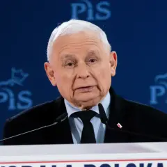 Jarosław Kaczyński w garniturze przemawia za mównicą na tle niebieskiego baneru z logo PiS.