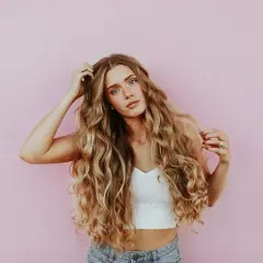 Ung kvinna med långt, vågigt blont hår som leker med håret framför en rosa vägg. Hon ser ut att ha sovit med inpackning.