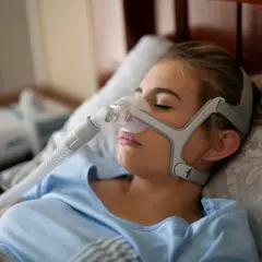 Junge Frau mit Schlafmaske, die gegen Schlafapnoe-Symptome hilft. Ein CPAP-Gerät ist im Hintergrund zu sehen.