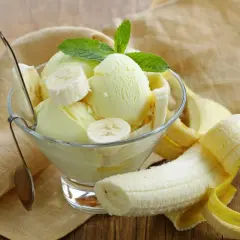 Pyszne lody bananowe w pucharku, ozdobione plasterkami banana i listkiem mięty. Obok leży obrany banan.