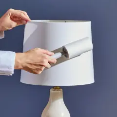 Hands using a lint roller to clean a white lampshade, demonstrating how to clean lamp shades effectively.