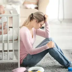 Zmęczona mama siedzi na podłodze obok łóżeczka, czując baby blues. Dziecko bawi się zabawką.