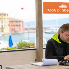 Uomo al lavoro con laptop, con vista sul porto e un cartello "Tassa di soggiorno".