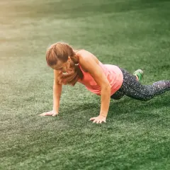 Une femme en tenue de sport fait des pompes sur une pelouse synthétique, un entraînement typique de boot camp sport.