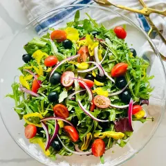 En fräsch italiensk sallad i en glasskål, fylld med gröna blad, körsbärstomater, svarta oliver, rödlök och gul paprika.
