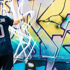 Mann mit Hut und Trikot sprüht mit Spraydose auf bunte Graffiti-Wand.