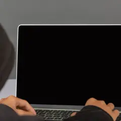 Osoba w kapturze pisze na laptopie z czarnym ekranem.