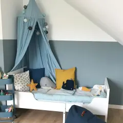 Ein Kinderzimmer, in dem blau und grau mischen ergibt eine beruhigende Wandfarbe. Ein Himmelbett mit Wal-Kissen und ein Teddybär auf einem Rollwagen.