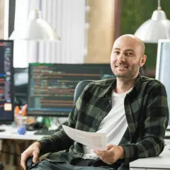 Ein Mann mit Glatze, der ein Dokument hält, sitzt vor mehreren Computermonitoren mit Code. Er könnte einen Techniker im Fernstudium bei der IHK absolvieren.