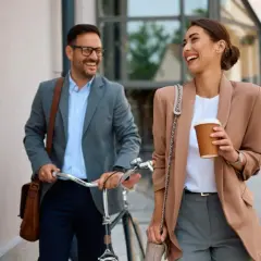 Uśmiechnięta para w strojach biznesowych idzie ulicą z rowerem i kawą.