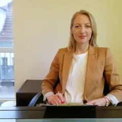 Kobieta w beżowej marynarce siedzi przy biurku i pisze.