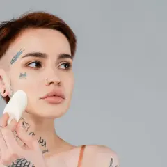 En kvinna med tatueringar applicerar foundation med en svamp för att jämna ut sin underton hud.