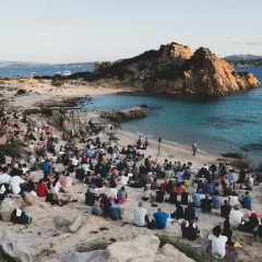 Concerto sulla spiaggia in Sardegna, con un pubblico seduto tra le rocce che ascolta un cantante sardo.