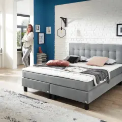 Modernes Boxspringbett in Grau, ideal für erholsamen Schlaf bei Bandscheibenvorfall. Eine Frau lehnt lässig daneben.