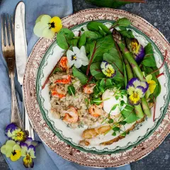A plate of creamy risotto with shrimp, asparagus, greens, and edible flowers, served with a fork and knife.