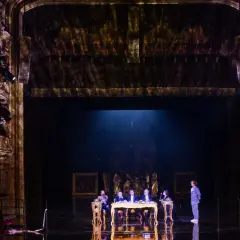 Na scenie teatru, przy złotym stole, siedzi pięć postaci. W tle widać bogato zdobione loże i kurtyny, tworząc atmosferę historycznego przedstawienia.
