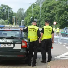 Służba celno skarbowa kontroluje ruch na drodze. Dwóch funkcjonariuszy w kamizelkach odblaskowych stoi przy oznakowanym radiowozie.
