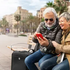Uśmiechnięta para seniorów sprawdza na telefonie, czy roaming jest domyślnie włączony w Plus, siedząc na ławce z walizką.
