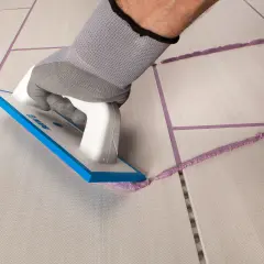 Mano con guanto grigio stende cemento e acqua senza sabbia con spatola su fughe di piastrelle.