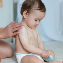 Mama smaruje kremem ramię dziecka, które siedzi w prawidłowym siadzie. Dziecko trzyma w rączce niebieską kostkę.