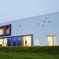 Nowoczesny, ocieplony dom bez ogrzewania, z dużymi przeszkleniami i nietypowymi okrągłymi oknami. Wewnątrz widać sylwetkę osoby.