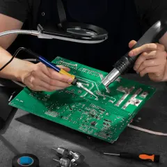 Ręce lutują płytę elektroniczną, używając lutownicy i gorącego powietrza. Widać też lupę, śrubokręty i taśmę izolacyjną – wszystko, co można lutować.
