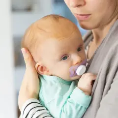 Malutkie dziecko z niebieskimi oczami i fioletowym smoczkiem dla niemowlaka w buzi, wtulone w ramiona mamy.