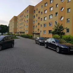 Osiedle Tysiąclecia Poznań. Jasnożółty blok z charakterystycznymi oknami w kształcie rombów, zaparkowane samochody na brukowanym podjeździe.
