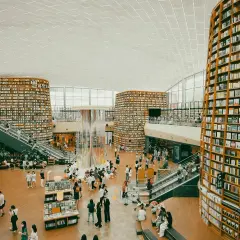 Wielopoziomowa biblioteka z półkami sięgającymi sufitu, gdzie każdy **bibliotekarz** marzyłby o pracy. Ludzie spacerują, czytają i odpoczywają wśród książek.
