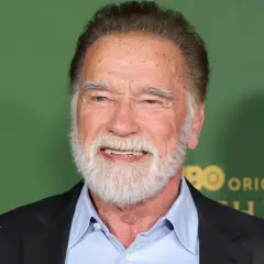 Arnold Schwarzenegger, z siwą brodą i w niebieskiej koszuli, uśmiecha się. W tle zielona ściana z logo HBO.