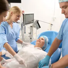 Uśmiechnięta pacjentka w czepku leży na łóżku, otoczona przez zespół medyczny. Opiekun medyczny i dwie pielęgniarki czuwają nad jej komfortem.